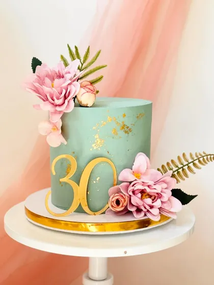 A modern and tropical cake in a lovely teal color. The cake is decorated with a large gold "30" topper, pink silk flowers, and faux fern leaves.