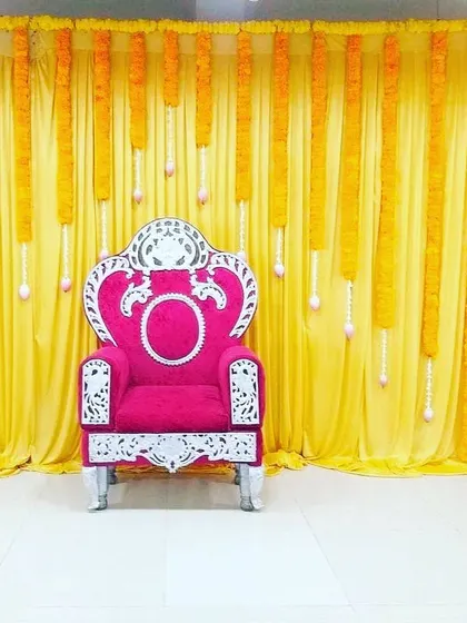 A simple and elegant backdrop featuring a bright yellow drape with hanging marigold garlands. This classic design is versatile and can be used for any small puja, Haldi ceremony, or festive gathering at home.