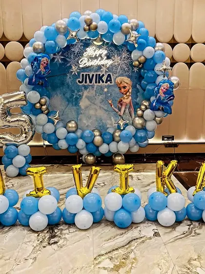 A magical Frozen-themed 5th birthday party. This grand setup includes a circular balloon frame in icy blue and silver, a custom backdrop featuring Elsa, and the birthday girl's name spelled out in foil balloons.