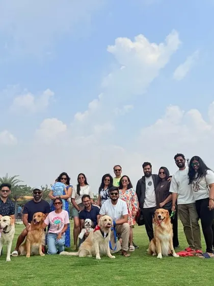 The whole squad goals picture! A large group of pet parents and their dogs gather for a photo on our lush green lawn under a clear blue sky.