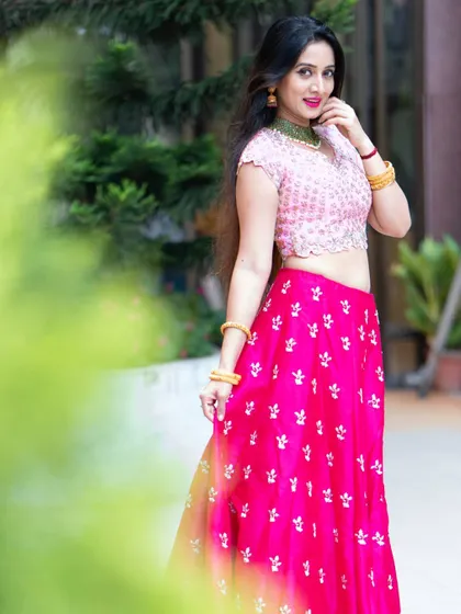 A vibrant outdoor portrait of actress Harshika Poonacha. The bright pink lehenga and her joyful expression create a lively and energetic photo.