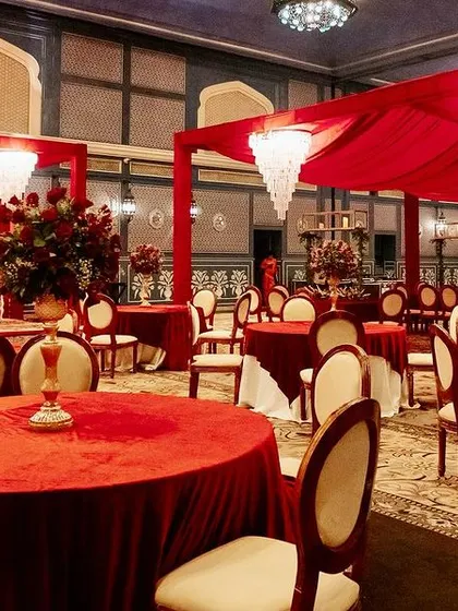 Guest dining tables set for a royal wedding, with red velvet tablecloths and elegant cream-colored chairs creating a rich and inviting look.