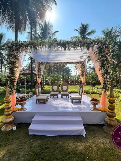 An elegant outdoor 'Muhurtham Mantap' set against a beautiful backdrop of palm trees. The simple white stage, pastel drapes, and fresh floral arrangements create a serene and picturesque setting for the wedding ceremony.