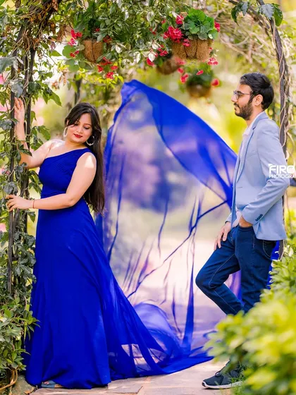 A candid, romantic moment in the garden. The groom-to-be looks on as his partner poses among the vines, with the flowing blue fabric of her gown catching the light beautifully.
