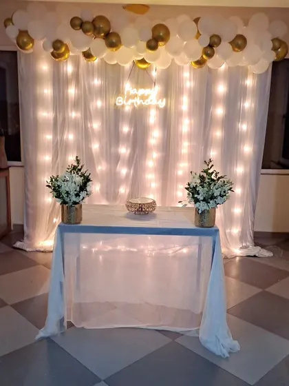 A simple and elegant decoration for a surprise birthday party. The backdrop of white curtains with fairy lights, combined with a white and gold balloon garland, creates a magical and intimate setting.