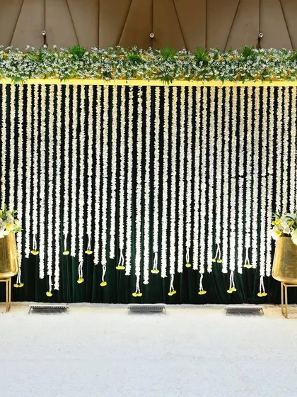 A clean and elegant Mehendi stage with a backdrop of white floral strings against dark green velvet. The gold accents in the vases add a touch of modern sophistication.