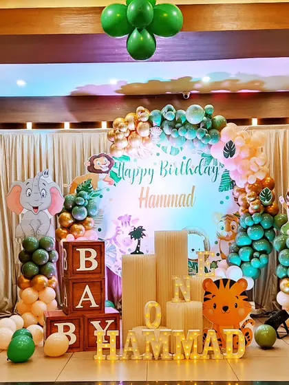 A cute baby jungle theme for a first birthday. This stage decoration includes a balloon arch in jungle greens and oranges, a picket fence with animal cutouts, and 'BABY' blocks for the perfect photo opportunity.