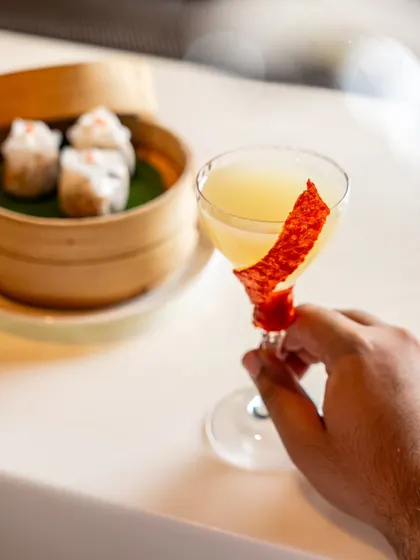 A hand holding a craft cocktail at Yautcha, with dim sum in the background. This image connects the drink to the dining experience.