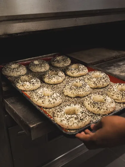 A tray of boiled bagels, generously topped with seeds, is loaded into the oven for the final bake.