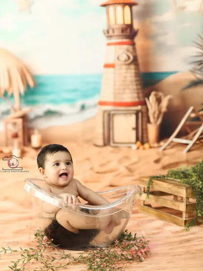 A happy baby enjoying a bath in front of a lighthouse. The detailed beach backdrop, complete with sand and greenery, makes for a very imaginative portrait.