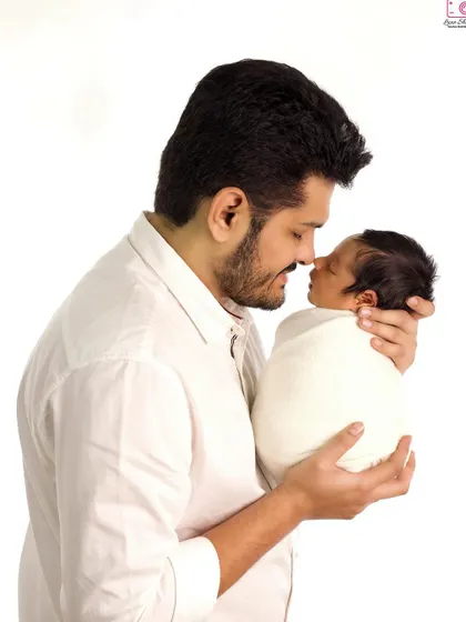 A classic portrait of a father holding his swaddled newborn, their faces close together. The clean white background emphasizes the emotional bond.
