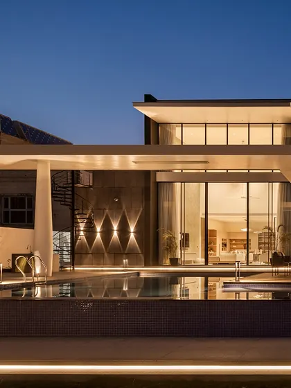 The back elevation of the Ananda residence at night. The architectural lighting highlights the clean lines, the inviting pool, and the seamless transition to the covered patio, creating a calm and magical atmosphere.