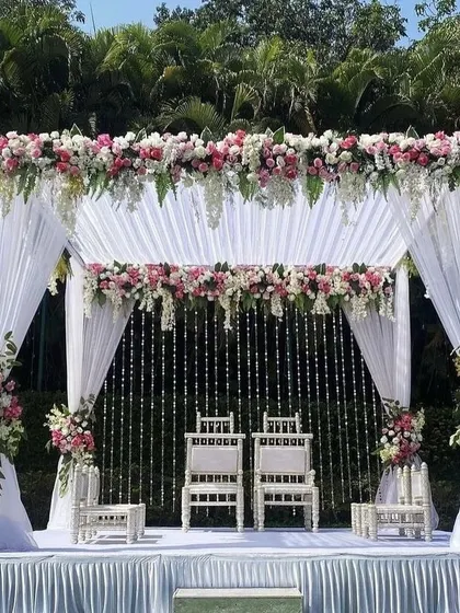 An elegant and classic outdoor wedding mandap design. This setup features a canopy of white drapes, lush pink and white floral arrangements, and delicate crystal hangings, creating a serene and beautiful space for the wedding ceremony.