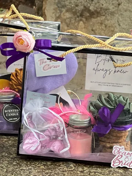 A bridesmaid proposal hamper displayed outdoors. The clear box contains a scented candle, cookies, and a card with a playful message asking, "Will you be my bridesmaid?".