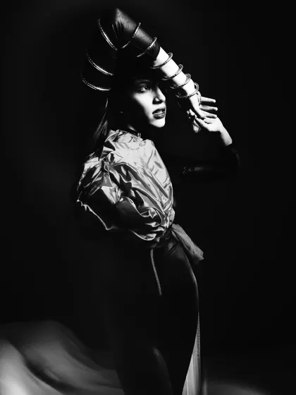 A dramatic black and white portrait using strong side lighting. The model's pose is statuesque, and the monochrome treatment emphasizes the play of light and shadow on her form and the sculptural headpiece.