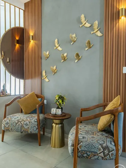 This cozy corner features patterned accent chairs against a fluted wood wall. The design is enhanced by a large round mirror and a flock of golden bird wall sculptures, adding a touch of whimsy and artistic flair.