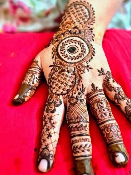 A detailed party henna design on the hand, featuring circular motifs, checkerboard patterns, and intricate finger details.