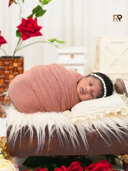 A newborn sleeps peacefully on a miniature wooden bed, wrapped in a soft brown swaddle. The scene is decorated with red and white roses, creating a romantic and gentle atmosphere.