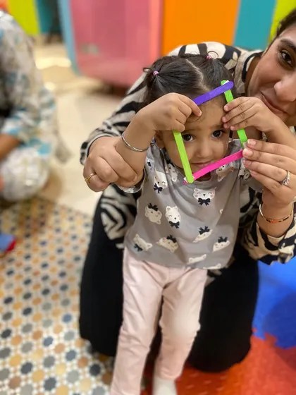 A toddler playfully peeks through a shape made from colorful sticks, combining learning with imaginative play.