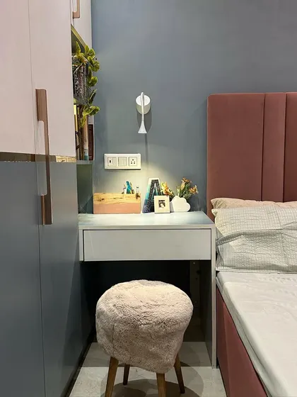 A cozy corner in a guest room, featuring a compact dressing table next to the bed. The pink upholstered headboard and fluffy stool add soft textures to the space.