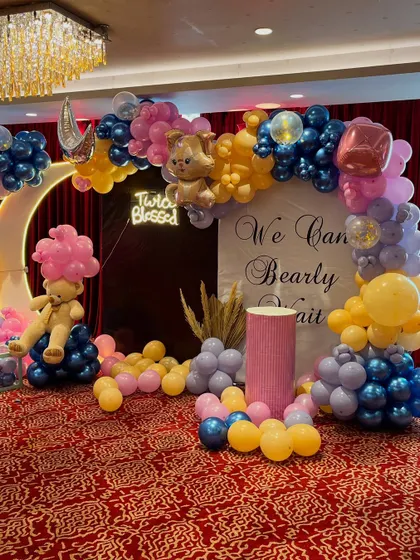 A full view of the twin baby shower decoration, capturing the entire balloon arch, the lighted moon, the neon sign, and the props, all set against a rich red curtain.