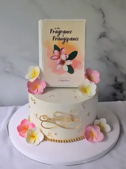 A beautiful and elegant cake designed for a book launch. The centerpiece is an edible replica of the book cover for "In the Fragrance of Frangipanis," surrounded by handcrafted sugar flowers that match the theme.