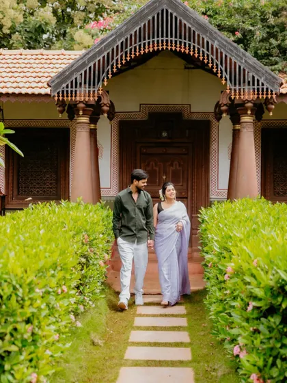 A couple dressed in beautiful ethnic wear, walking hand-in-hand down a path towards a traditional Indian-style home.