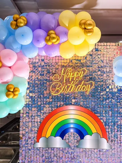 A 'Happy Birthday' sign on a silver sequin wall, framed by a pastel rainbow balloon arch.