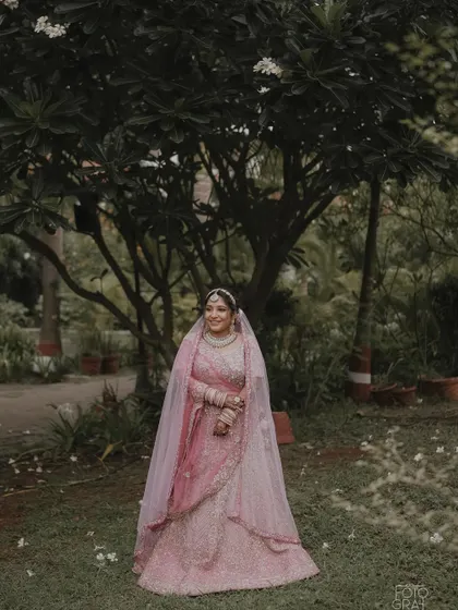 A bride in a beautiful pink lehenga stands in a garden, her long veil creating a soft, ethereal frame around her.