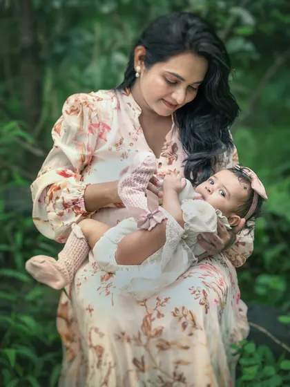 A mother gazes lovingly at her baby girl resting in her lap during an outdoor photoshoot. The soft lighting and lush green surroundings create a peaceful and intimate portrait of motherhood.