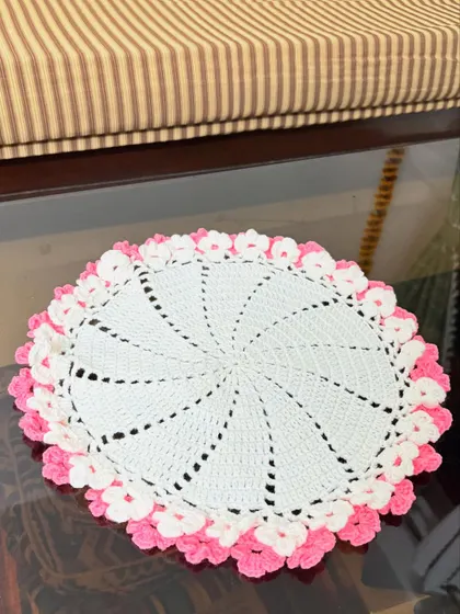 A delicate white crochet doily with a swirling pinwheel pattern and a lovely pink floral border.