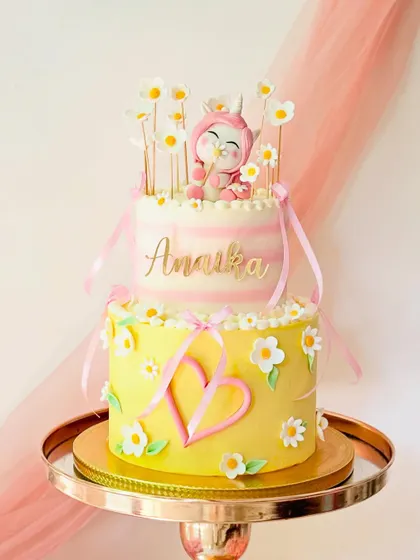 A cheerful two-tiered cake with a bunny-fairy character on top. The design uses bright yellow and pink, with daisy and heart details.