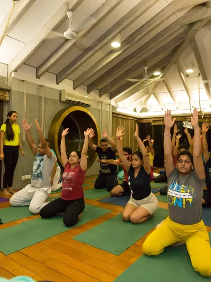 A class practices Virasana (Hero Pose) with arms extended overhead. This pose stretches the thighs and ankles while the arm variation opens the shoulders and chest.