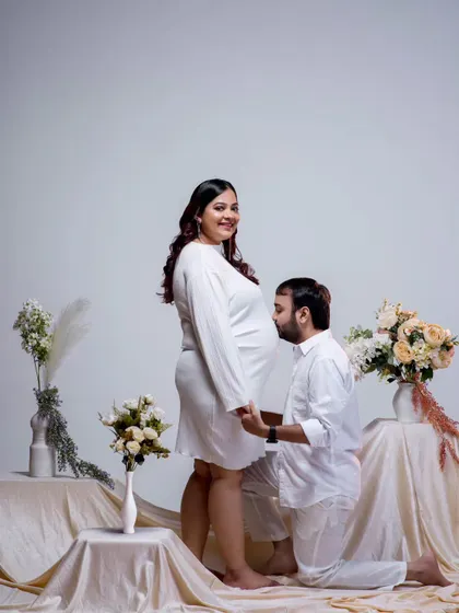 A classic pose filled with anticipation and love. The father-to-be kisses the baby bump, a gesture of affection for both mother and child. The all-white theme and floral arrangement create a pure and elegant setting.