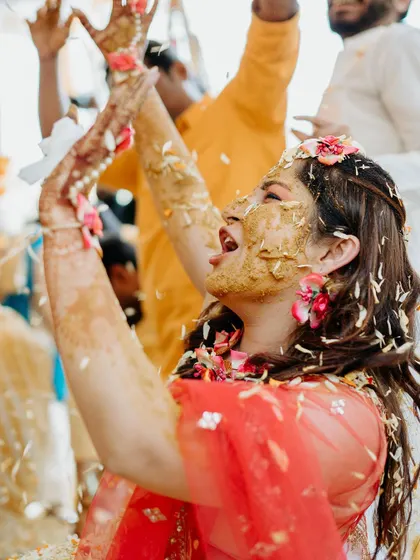 The bride throws her hands up in the air in sheer joy, covered in turmeric and flower petals during her Haldi.