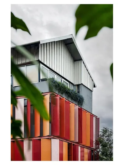 A view of the building with the red kinetic facade, framed by foliage. The design shows how a contemporary, industrial building can coexist with nature.