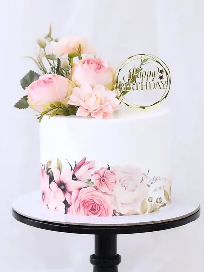 A pretty and elegant birthday cake featuring a printed floral wrap on the bottom tier. The design is topped with a delicate arrangement of fresh pink flowers and a "Happy Birthday" topper.
