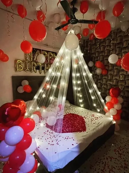 A cozy and romantic birthday setup with a lighted canopy over the bed, red and white balloons, and scattered rose petals.