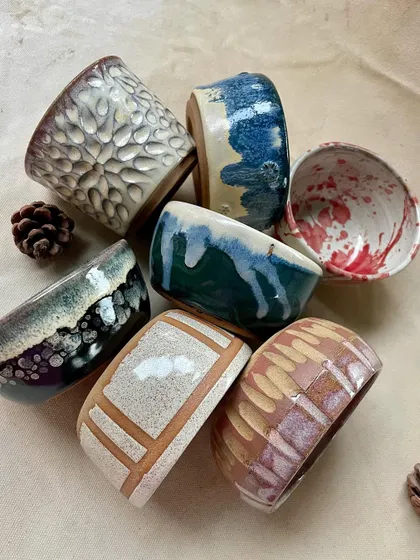 A collection of glazed student work, showing a range of styles and techniques. The pieces include a textured white cup, a speckled bowl, and a mug with a carved house design.