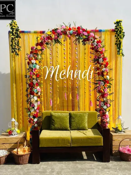 A bright and cheerful Mehndi backdrop. I used a vibrant yellow curtain as the base, adorned with a multi-colored floral arch and a simple 'Mehndi' sign for an elegant look.