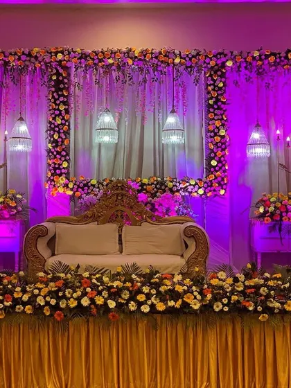 An elegant wedding stage design with a beautiful floral frame and dramatic purple uplighting. The combination of hanging birdcage lamps, candle stands, and a plush sofa creates a sophisticated and grand backdrop for the couple.
