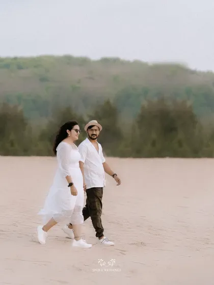The couple taking a leisurely walk on a wide, sandy beach, with hills in the background.