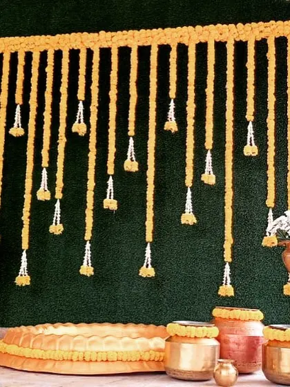 This is a classic haldi ceremony setup. The bright marigold flower frame and hanging garlands against a green backdrop, along with traditional brass and copper pots, create the perfect vibrant setting for the ritual.