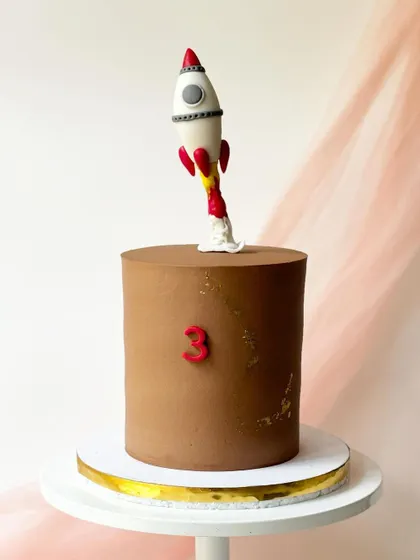 A minimalist rocket cake. The simple chocolate buttercream provides a clean backdrop for the fondant rocket topper, which is sculpted to look like it's blasting off.