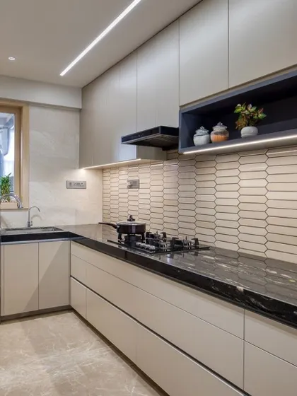 A sleek L-shaped kitchen with beige handleless cabinets and a black granite countertop. The open shelving unit with integrated lighting is perfect for displaying decor or frequently used items.