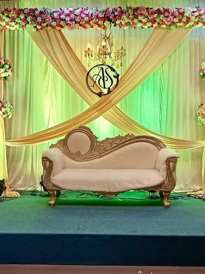 This engagement stage combines gold fabric draping with green backlighting for a classic look. The setup is completed with a central monogram, a chandelier, and floral bouquets on stands.