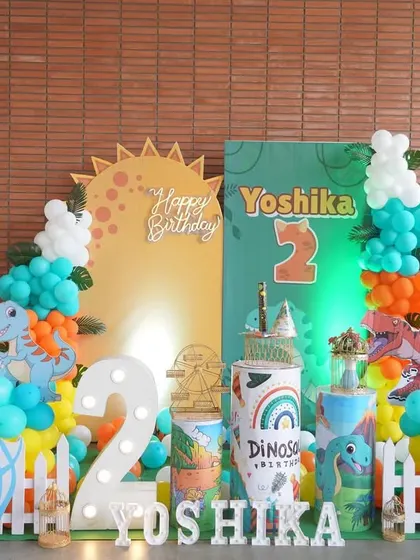 A roaring good time with this dinosaur-themed second birthday party. The decor includes a variety of dinosaur cutouts, a volcano backdrop, and a light-up number '2' surrounded by a white picket fence.