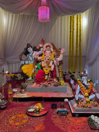 A full view of the pandal setup for the 'Gokul Nagari Cha Maharaja' 2024 event. This shows the simple yet clean arrangement with white fabric draping, a pink chandelier, and a well-lit space for multiple idols.