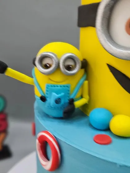 A close-up of a playful Minion peeking out from the side of a colorful two-tier cake.