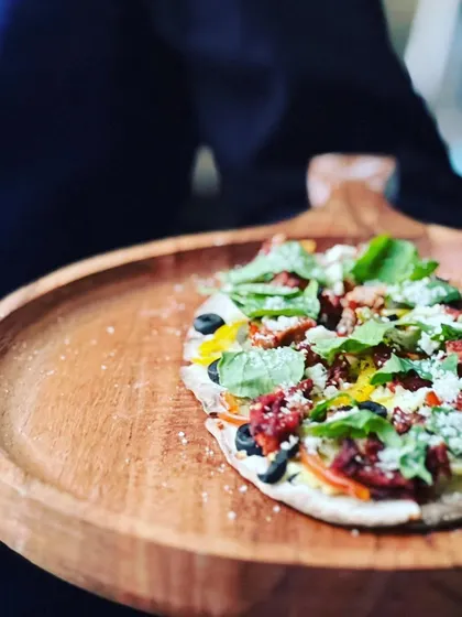 A freshly baked flatbread pizza, topped with sun-dried tomatoes, olives, and fresh basil, served on a rustic wooden board.